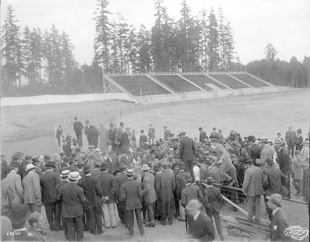 Schwarzes und weißes Foto einer Menge, die sich um ein Fussballfeld versammelt hat, während der Eröffnungszeremonien der Olympischen Spiele 1904, mit Bäumen und einem klaren Himmel im Hintergrund.