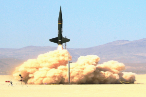 Eine Rakete startet vom Boden aus und steigt in den Himmel auf, hinter sich eine Rauchspur, vor einer Bergkulisse.