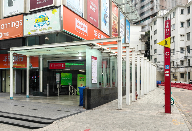 Eine Stadtstraße mit einem Bushaltestellen, Gebäude auf beiden Seiten, eine Straße mit einem Müllcontainer, rote und gelbe Schilder mit Text, Menschen auf dem Gehweg und eine Rolltreppe auf der linken Seite.