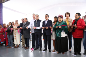 Eine Gruppe von Menschen bei einer Eröffnungszeremonie mit einer Schere, einer Säule links daneben, verstreuten Papieren auf dem Boden und einem weißen Tuch im Hintergrund.