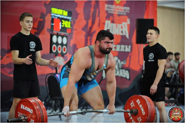 Ein Mann hebt eine Hantel bei einem Gewichtheben-Wettbewerb, umgeben von Zuschauern, mit Wettbewerbsequipment und Beschilderung, die die 2018 World Weightlifting Championships anzeigt.