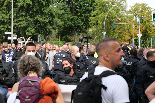 Eine große Gruppe von Menschen, einige mit Masken und Rucksäcken oder Kameras, steht vor einer Menge von Polizeibeamten, mit Laternenmasten, Ampeln, Verkehrszeichen, Bäumen und einem klaren blauen Himmel im Hintergrund.