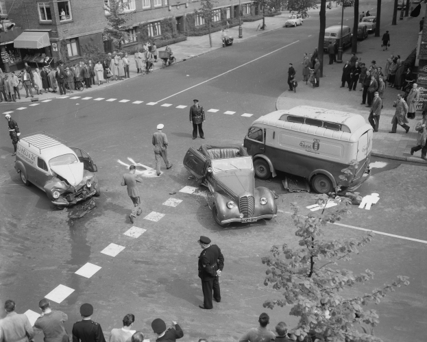 Eine schwarz-weiße Szenerie eines Autounfalls an einer Straßenkreuzung mit mehreren Fahrzeugen, darunter ein Lieferwagen, und einer Gruppe von Menschen drumherum, mit Gebäuden, Bäumen, Pfählen und Texttafeln im Hintergrund.