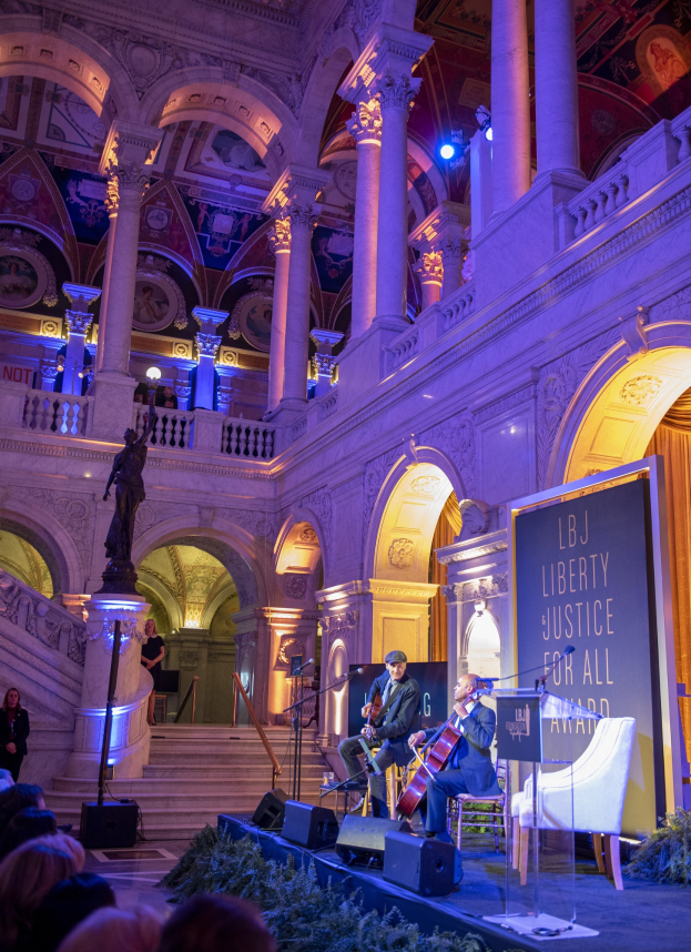 A group of people seated on a stage during the Liberty Justice for All Award Ceremony in New York City, with musicians performing, a podium with a microphone, a board with text, and architectural elements including a statue, pillars, and ceiling lights.