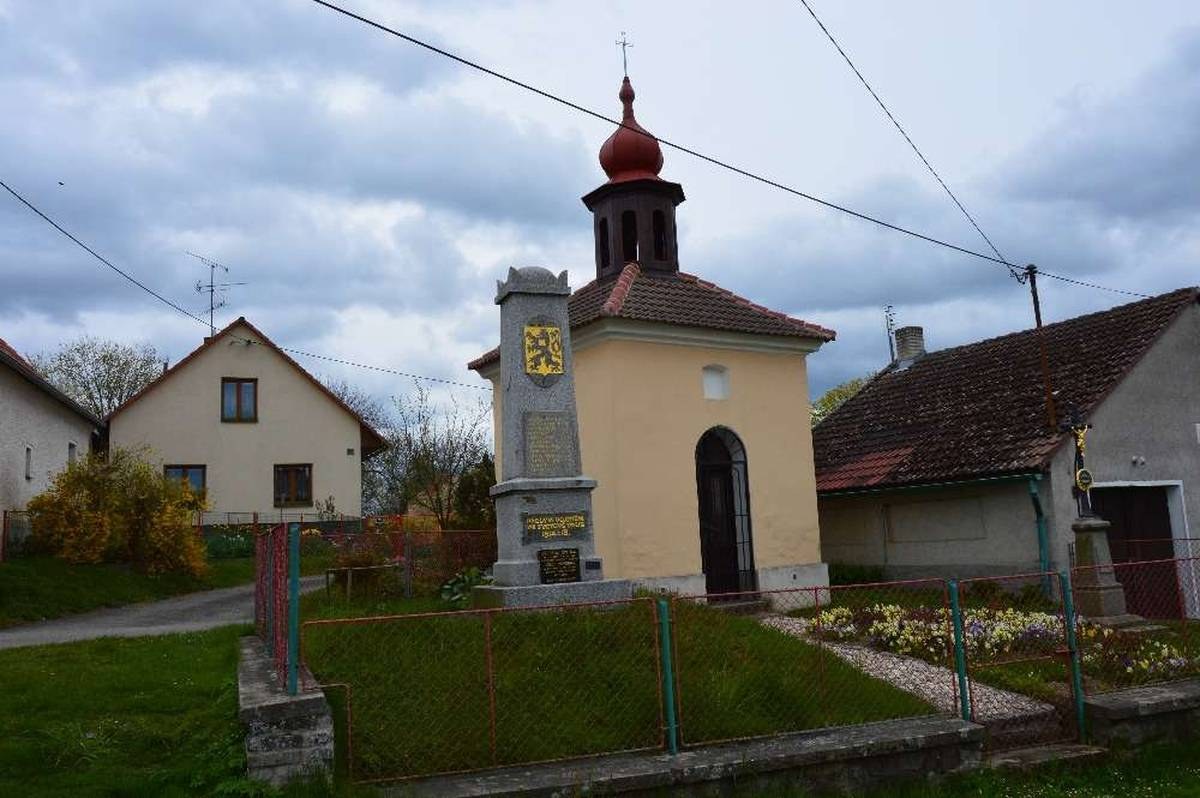 Kleine Kirche mit einem Glockenturm, identifiziert als die Kirche des Heiligen Grabes, umgeben von Häusern, Grünflächen, einem Zaun und einem bewölkten Himmel.