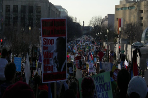 Eine Gruppe von Menschen, die auf einer Straße protestieren und Plakate halten, mit Gebäuden, Bäumen, Laternenpfählen und dem Himmel im Hintergrund.