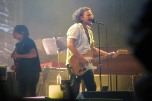 Ein Mann in einem weißen Hemd spielt Gitarre und singt in ein Mikrofon auf der Bühne, neben ihm spielt ein Mann in einem schwarzen Hemd Gitarre; ein Piano, Kabel, Lautsprecher und Lichter sind im Hintergrund zu sehen.