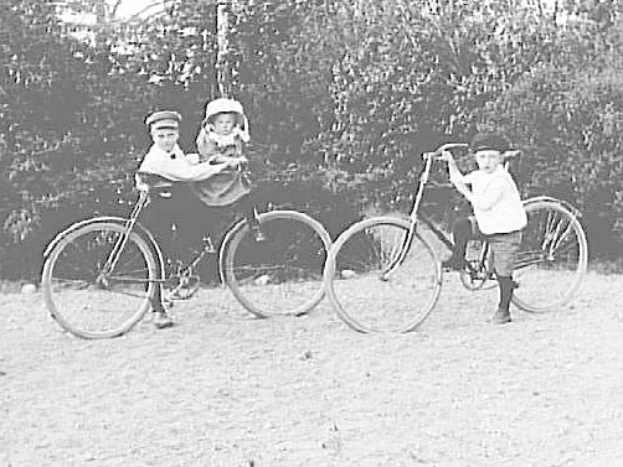 Zwei Kinder fahren Fahrräder in einem Park, eines hält ein Baby, mit Bäumen und einem Pfahl im Hintergrund, alles in Schwarz-Weiß.