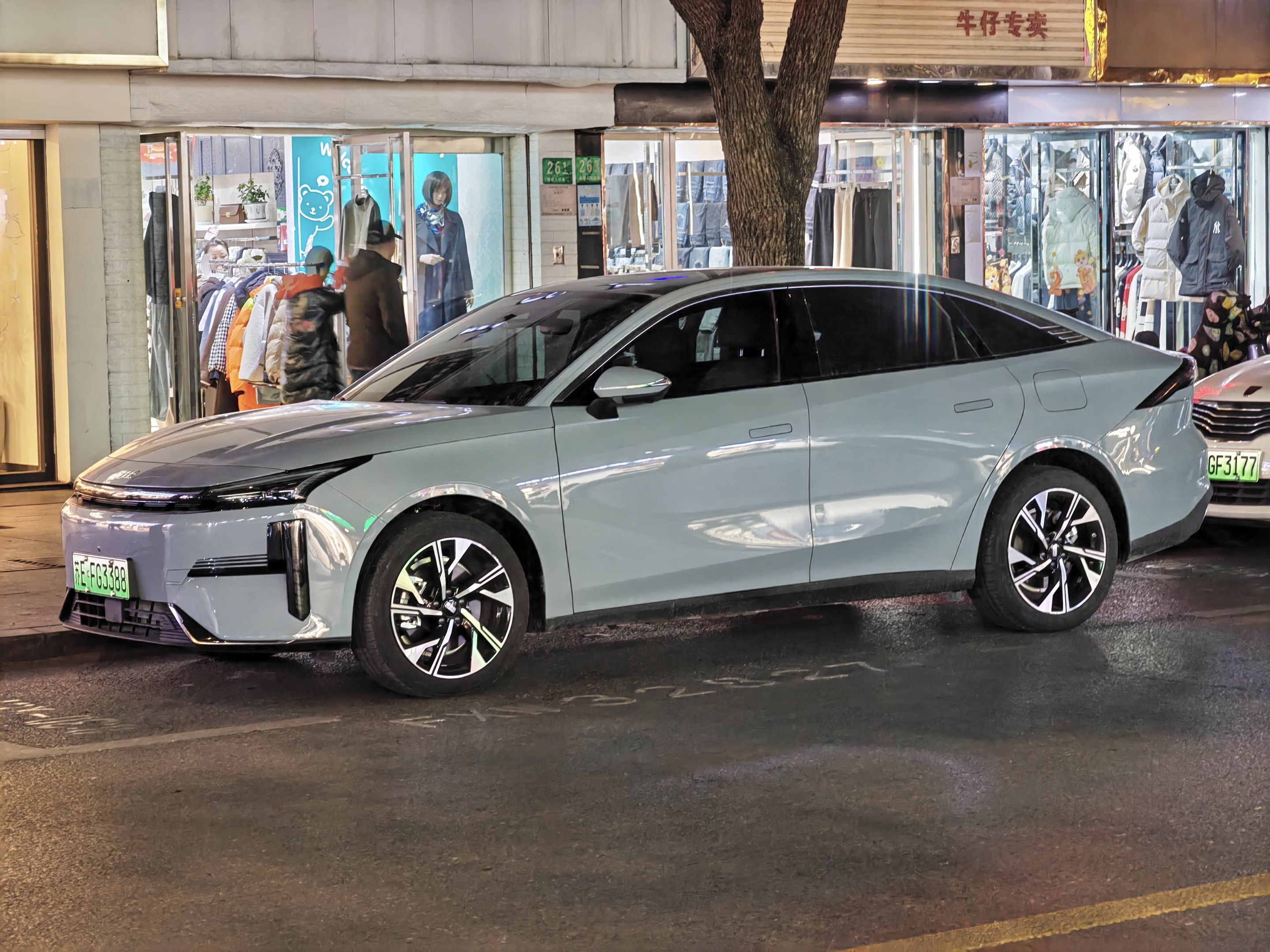 Elektrischer Mitsubishi Lancer EV auf einer nächtlichen Stadtstraße geparkt, umgeben von beleuchteten Schaufenstern, Schaufensterpuppen in Kleidern und Beschriftungen.