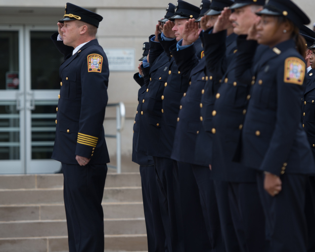 Eine Gruppe von Polizeibeamten in Uniform und Mützen, die in Formation vor einem Glastür-Gebäude salutieren.
