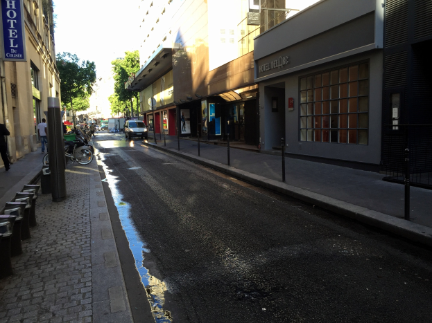 Eine Stadtstraße mit einer Pfütze am Straßenrand, fahrende Fahrzeuge, Fußgänger auf dem Gehweg, Gebäude mit Namensschildern, Bäume und einen klaren blauen Himmel im Hintergrund.
