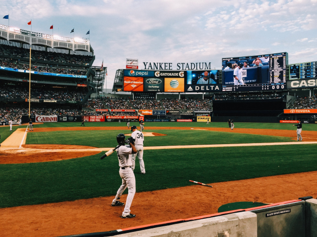 Baseballspiel im Gange im Yankee Stadium mit Spielern und Zuschauern, umgeben von Stadionmerkmalen wie ZÀunen, Flaggen, Anzeigetafeln, einem Anzeigebildschirm und Deckenleuchten unter einem bewölkten Himmel.