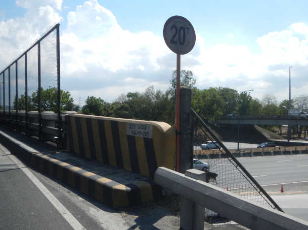 Geschwindigkeitsbegrenzungsschild auf einer Brücke mit Fahrzeugen, einem Zaun, Pfählen, Lichtern, Bäumen und einem bewölkten Himmel im Hintergrund.