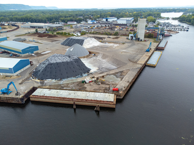 Luftaufnahme eines großen Gewässers mit einem zentralen Dock, umgeben von Gebäuden, Schuppen, Fahrzeugen, Kränen und anderen Gegenständen, mit Bäumen, Hügeln und einem klaren blauen Himmel im Hintergrund.