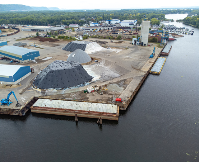 Luftaufnahme eines großen Gewässers mit einem zentralen Dock, umgeben von Gebäuden, Schuppen, Fahrzeugen, Kränen und anderen Gegenständen, mit Bäumen, Hügeln und einem klaren blauen Himmel im Hintergrund.