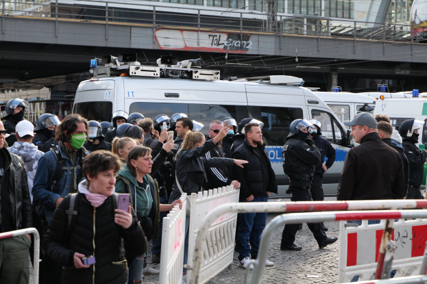 Eine Gruppe von Menschen vor Polizeifahrzeugen mit Absperrungen im Vordergrund und einer Brücke im Hintergrund während einer Demonstration in Berlin, Deutschland.