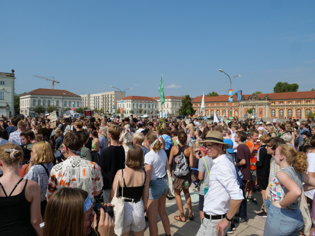 Große Menschenmenge vor einem Gebäude mit Fahnen, Bäumen und einem bewölkten Himmel, einige halten Kameras.