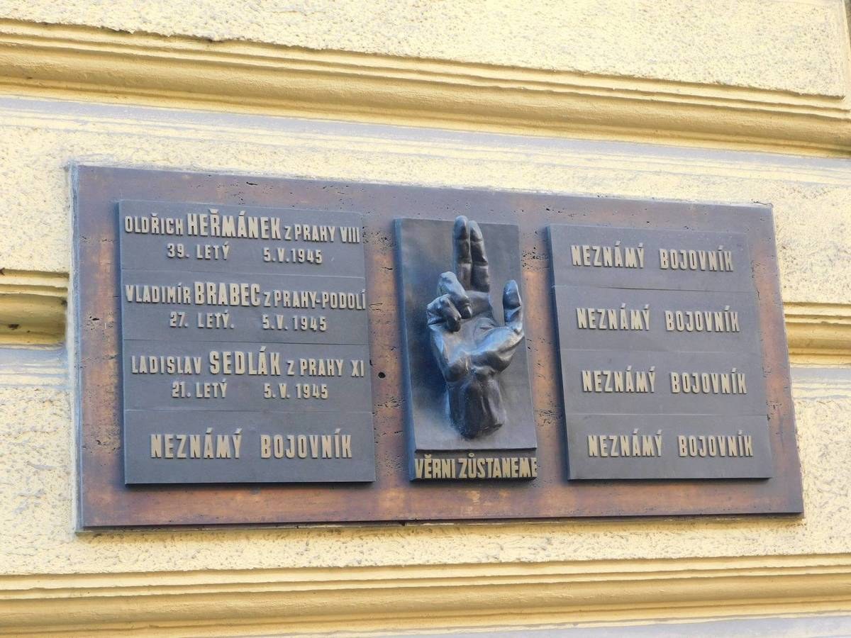 Metal plaque on a building in Prague, Czech Republic, with engraved text and a sculpted hand.