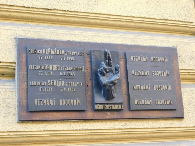 Metal plaque on a building in Prague, Czech Republic, with engraved text and a sculpted hand.