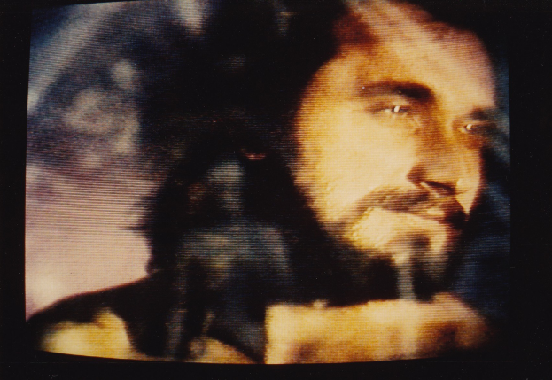 Ein Mann mit langem dunklem Bart und Schnurrbart, der ein weißes Hemd trägt, erscheint auf einem Fernseher mit dunklem Hintergrund und schaut direkt in die Kamera mit einem ernsten Gesichtsausdruck.