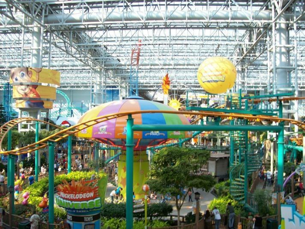 Großes Indoor-Wasserpark mit Achterbahn, umgeben von Menschen, Bäumen, Pflanzen und Deckenbeleuchtung, die eine lebendige Atmosphäre schaffen.