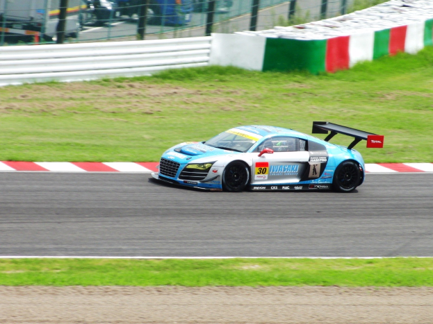 Audi R8 LMS GT3 Rennwagen auf einer Rennstrecke mit Gras auf beiden Seiten, umgeben von einem unscharfen Hintergrund einschließlich eines Zauns und Fahrzeugen.