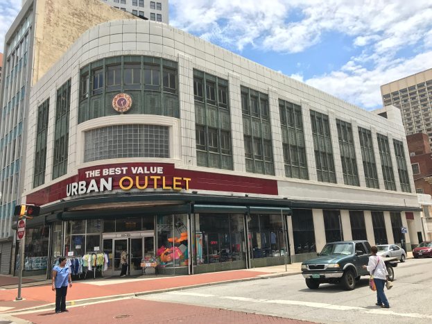 Eine belebte Stadtstraße mit Fahrzeugen auf der Straße und Fußgängern auf dem Gehweg, mit hohen Gebäuden mit Glasfenstern und einem Schild mit der Aufschrift "Der beste Preis für die Stadt" im Hintergrund unter einem bewölkten Himmel.