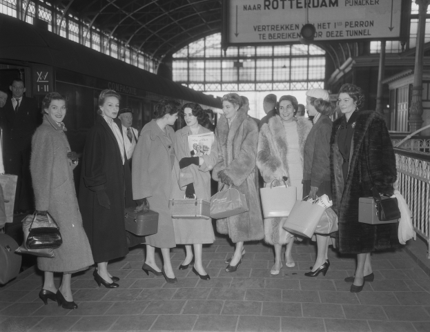 Black and white image of a group of women standing together at a train station, each holding a bag, with a train on the left and railings and a board on the right.