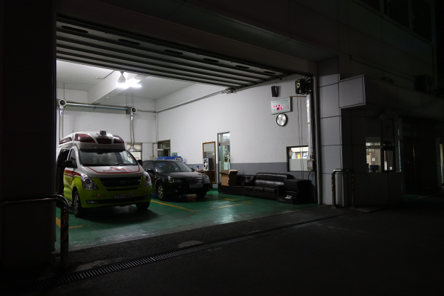 Ambulanz in einem Gebäude bei Nacht geparkt, mit sichtbaren Fahrzeugen, Sofas, einer Wanduhr, einer Texttafel, einer Tür, Rohren, Deckenleuchten und Metallstangen.