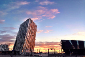 Eine Stadtansicht mit einem prominenten Wolkenkratzer, mehreren anderen Gebäuden, einer Werbetafel auf der rechten Seite, einer Straße mit einem Auto unten und Wolken am Himmel darüber.