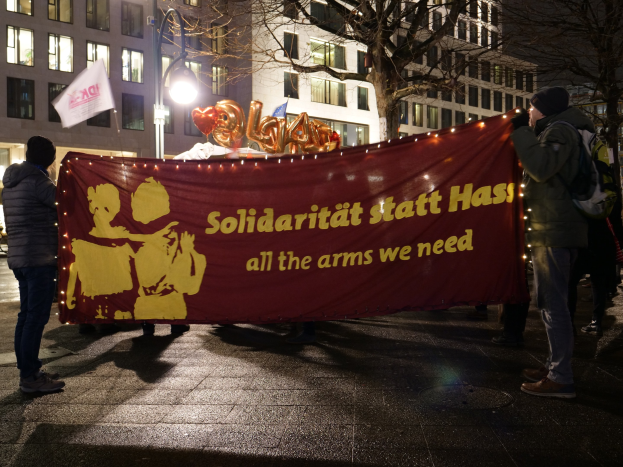 Gruppe von Menschen mit einer Tafel "Solidarität statt Hass - alle Waffen, die wir benötigen" im Support der Holocaust, mit Fahrzeugen, Bäumen, Gebäuden und einem bewölktem Himmel im Hintergrund.