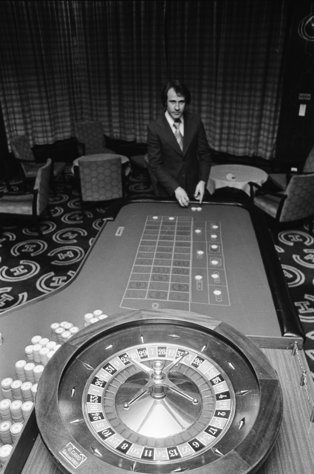 Schwarzes und weißes Bild eines Mannes in einem Anzug, der an einem Roulettetisch in einem Casino sitzt und von Stühlen und Tischen umgeben ist, mit einem Vorhang im Hintergrund.