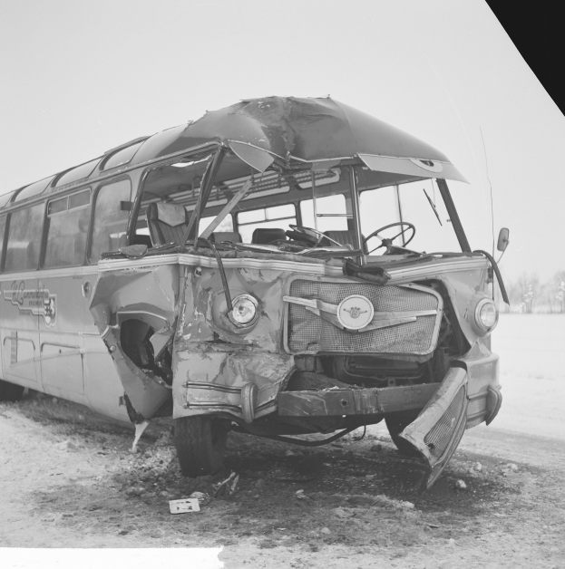Schwarz-weiß-Foto eines schwerbeschädigten Busses mit eingedrütkter Front und zersplitterten Fenstern am Röstrand, umgeben von Bäumen unter einem bewölktem Himmel.