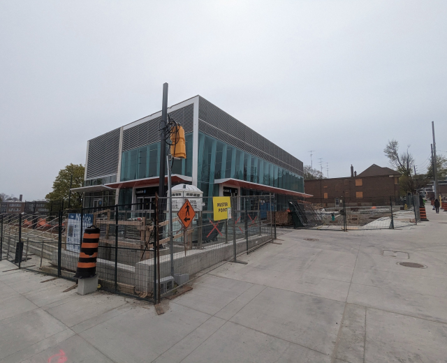 Large multi-story school building with numerous windows, surrounded by a metal fence, trees, signboards, and a few people and vehicles; cloudy sky overhead.