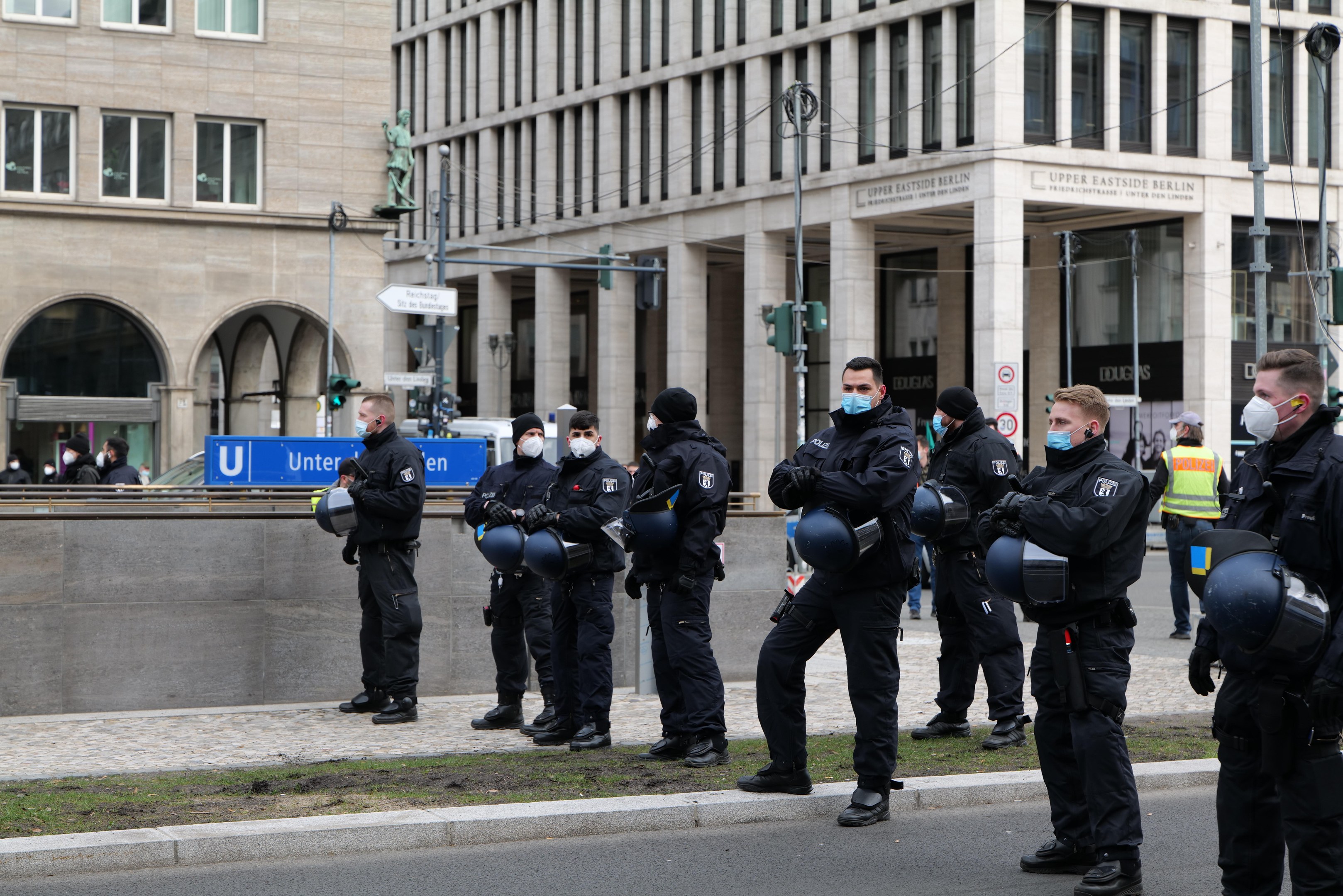 Polizisten in schwarzen Uniformen und Masken vor einem Gebäude mit Glasfenstern und Säulen in Berlin, Deutschland, einige mit Helmen, mit Laternen, Verkehrszeichen, Schildern mit Text, einer Statue und anderen Menschen im Hintergrund.