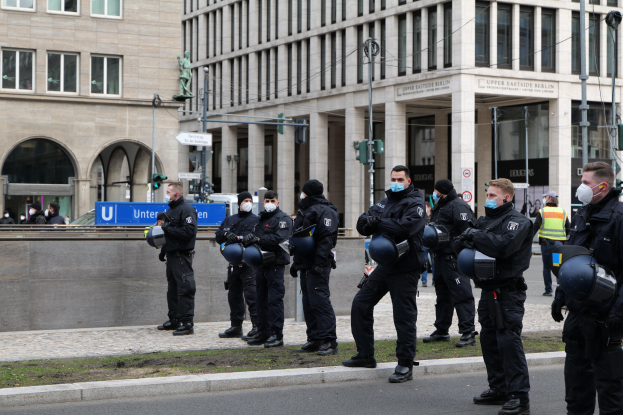 Polizisten in schwarzen Uniformen und Masken vor einem Gebäude mit Glasfenstern und Säulen in Berlin, Deutschland, einige mit Helmen, mit Laternen, Verkehrszeichen, Schildern mit Text, einer Statue und anderen Menschen im Hintergrund.
