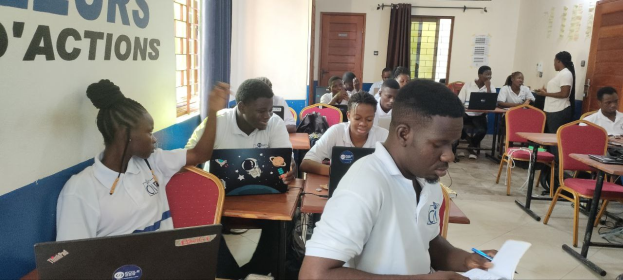 Eine Klasse mit Schülern an Tischen, die Laptops und Bücher verwenden, mit Unterrichtsmaterialien an Wänden und Fenstern, was auf ein E-Learning-Programm in Ghana hinweist.