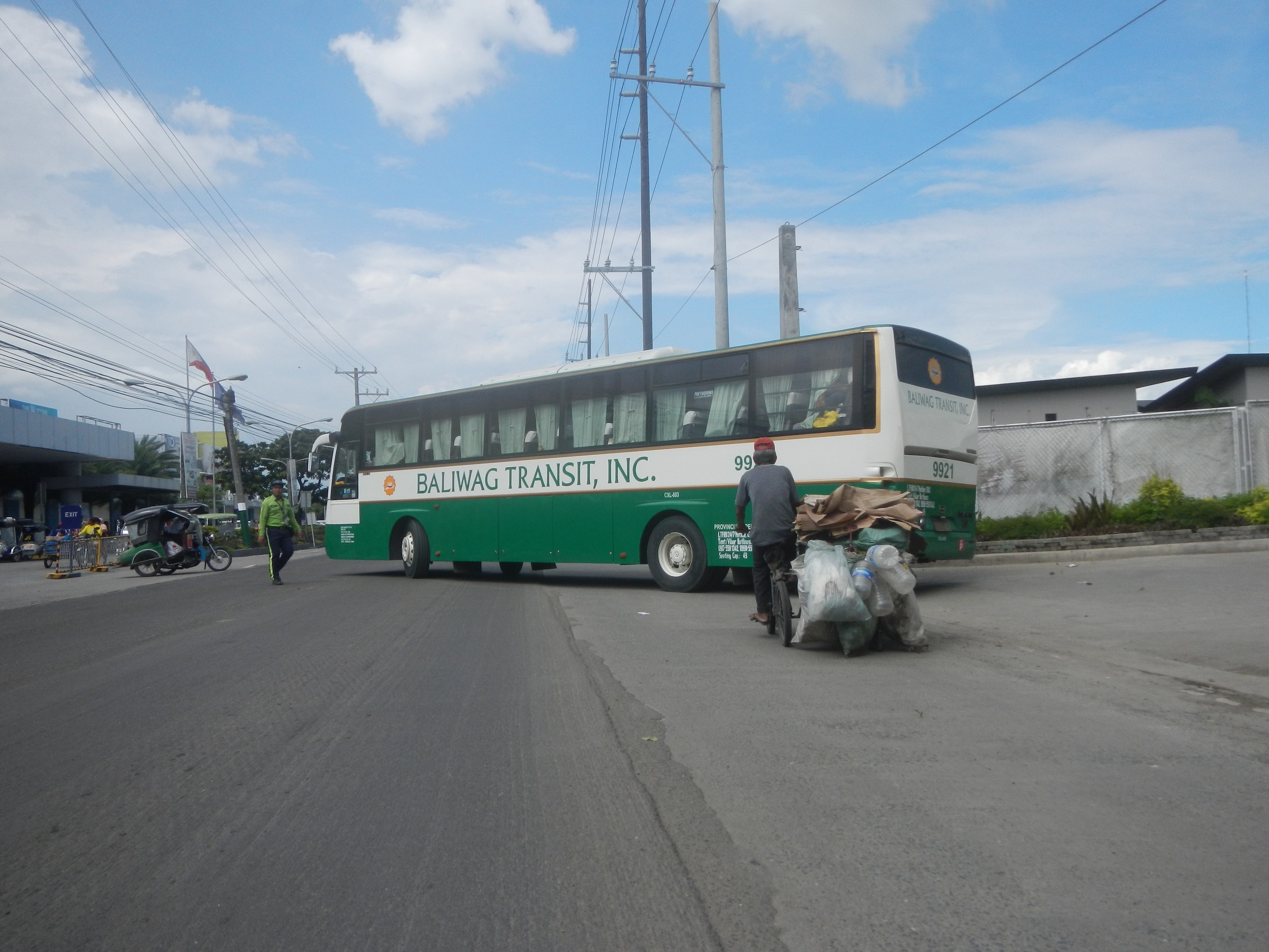 Ein grüner und weißer Bus fährt auf einer Straße neben einem Gebäude, vorbei an Fußgängern, einem Radfahrer, Strommasten, Gebäuden, Bäumen und einem bewölkten Himmel.