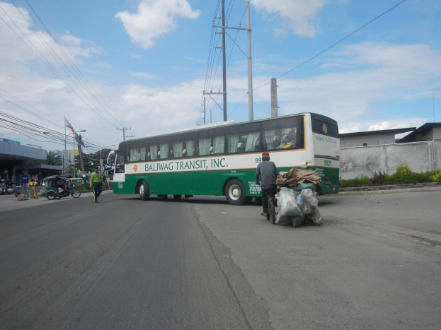 Ein grüner und weißer Bus fährt auf einer Straße neben einem Gebäude, vorbei an Fußgängern, einem Radfahrer, Strommasten, Gebäuden, Bäumen und einem bewölkten Himmel.