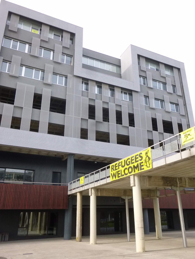 Ein großes Gebäude mit einem "Flüchtlinge Willkommen"-Schild, flankiert von Säulen und einem Geländer, unter einem sichtbaren Himmel.