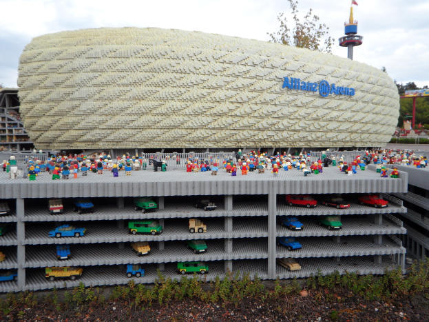 A large Lego model of the Allianz Arena in Munich, Germany, with people standing in front, surrounded by trees, poles, a tower, and plants, under a cloudy sky.
