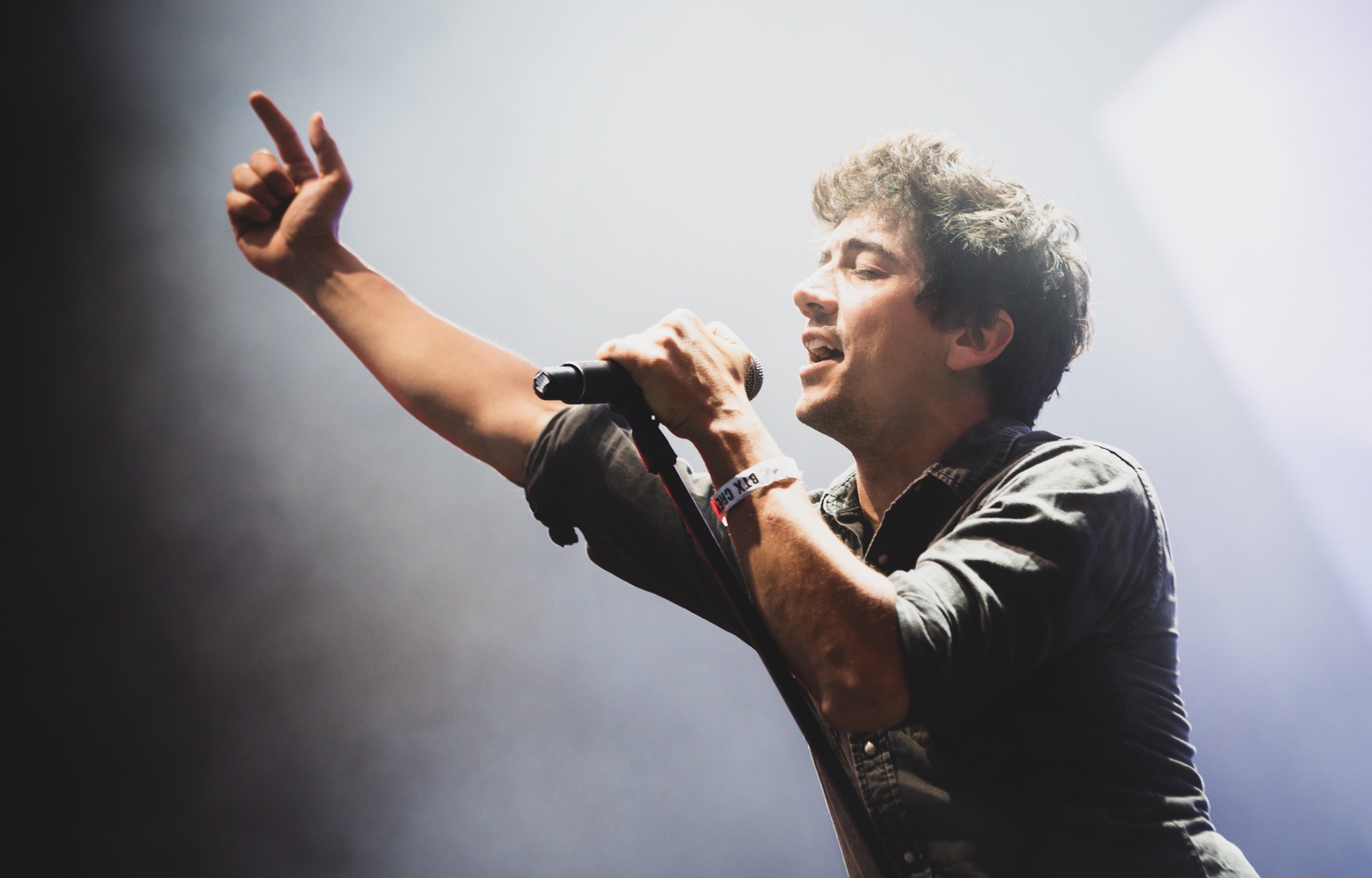 A man in a black shirt and jeans sings passionately into a microphone on a brightly lit stage, his eyes closed, as an applauding audience watches.