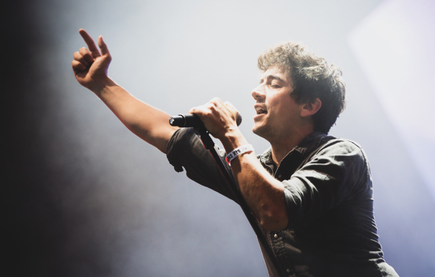 A man in a black shirt and jeans sings passionately into a microphone on a brightly lit stage, his eyes closed, as an applauding audience watches.