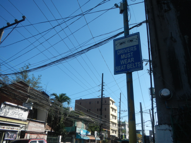 Stadtstraße mit fahrenden Autos, Strommöllen mit Drähten, Gebäude, Bäume und Namenschilder, mit einem "Fahrer müssen Sitzgürtel tragen"-Schild an einem Strommast gegen einen sichtbaren Himmel.