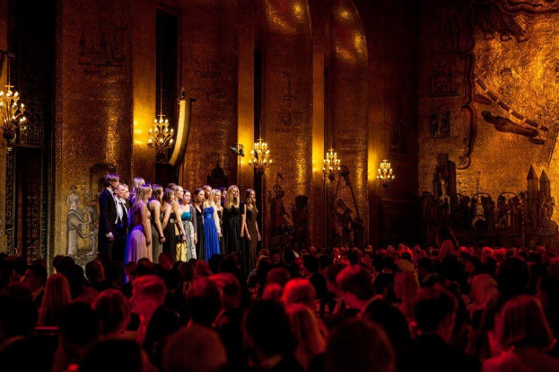 Eine Gruppe von Menschen auf einer Bühne im Royal Opera House in London, mit einem Publikum darunter, unter Bühnenbeleuchtung und umgeben von bemalten Wanddekorationen.