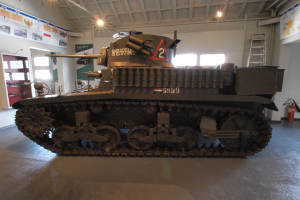 Ein großes britisches Armee-Panzerfahrzeug aus dem Zweiten Weltkrieg in einem Museum ausgestellt, umgeben von einem Feuerlöscher, einer Leiter, einem Feuerhydranten, einer Informationswand, einer Tür, Fenstern und Deckenleuchten.