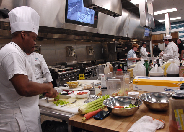 Eine Gruppe von Köchen in einer Küche, die Essen auf einem Schneidbrett zubereiten, wobei ein Koch in der foreground in einer weißen Schürze und einer Mütze steht und ein Messer hält und von Utensilien, Schüsseln, Gemüse, einem Mobiltelefon, Flaschen und anderen Gegenständen umgeben ist, mit einem Fernseher und Deckenlampen im Hintergrund.