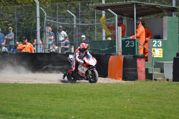 Ein Motorradfahrer in Helm und Handschuhen fährt über ein grasbewachsenes Feld, mit Zuschauern in Mützen und Schutzbrillen, Maschendrahtzaun, Treppen mit Geländern und einem nummerierten Schuppen im Hintergrund.