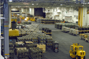 Ein großes Lagerhaus voller Numerous Trucks, Container, Menschen, Lichter, Stangen und andere Gegenstände, mit einem prominenten Rad.