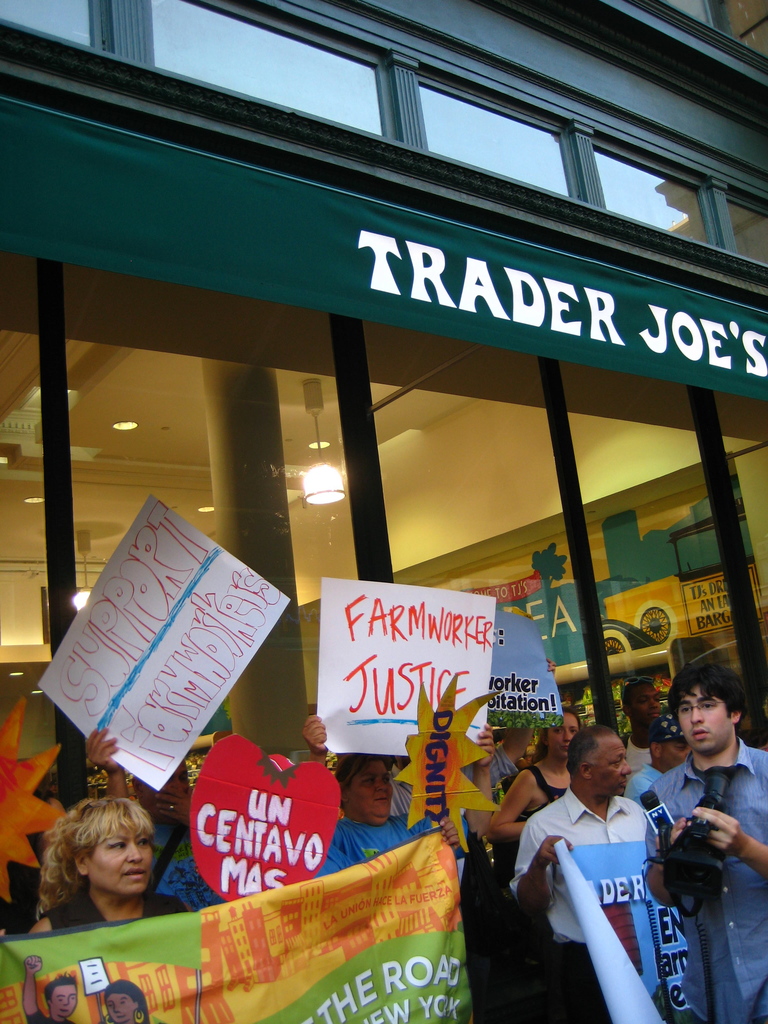 Eine Gruppe von Menschen steht vor einem Geschäft und hält Schilder und Plakate hoch, mit einer Säule und Laternen auf dem Dach im Hintergrund.